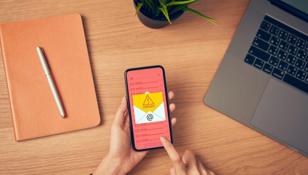Suspicious email warning on smartphone with malware alert, over wooden desk with laptop, notepad, pen, and plant, representing cybersecurity threats and email security solutions.