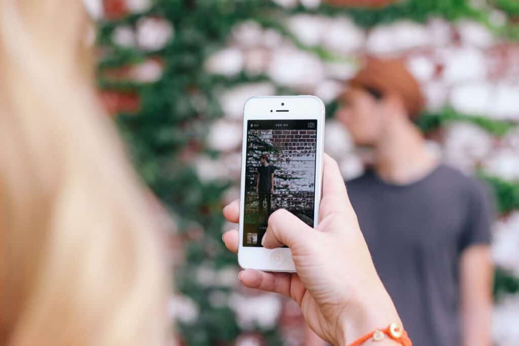 A person taking a photo of a woman with a smartphone outdoors against a brick wall.