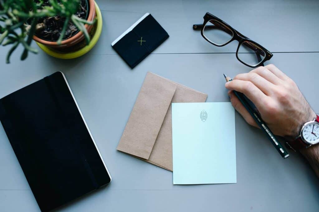 A person's hand holding a pen over stationery items, including a notebook, envelopes, glasses, and a potted plant on a gray desk surface, representing professional office or creative workspace.