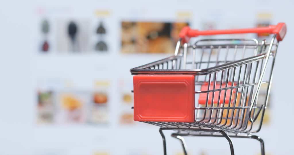 Miniature shopping cart on a white background, focused on the red front panel, with blurred store items in the background, symbolizing online shopping or retail services.