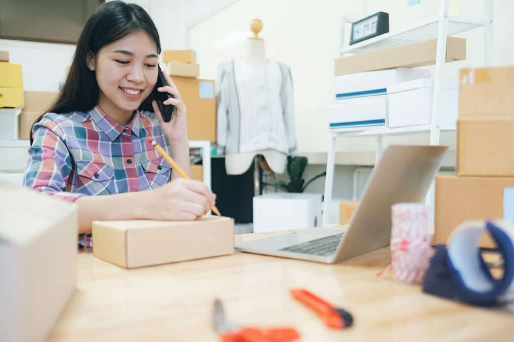 Young woman working in a small business or craft workshop, speaking on phone, taking notes with laptop and packing boxes.