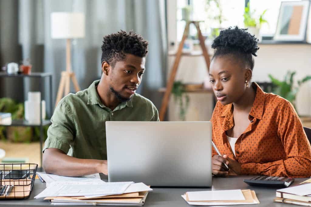 Young man and woman collaborating on a laptop in a modern office setting, discussing a project, surrounded by documents and office supplies.