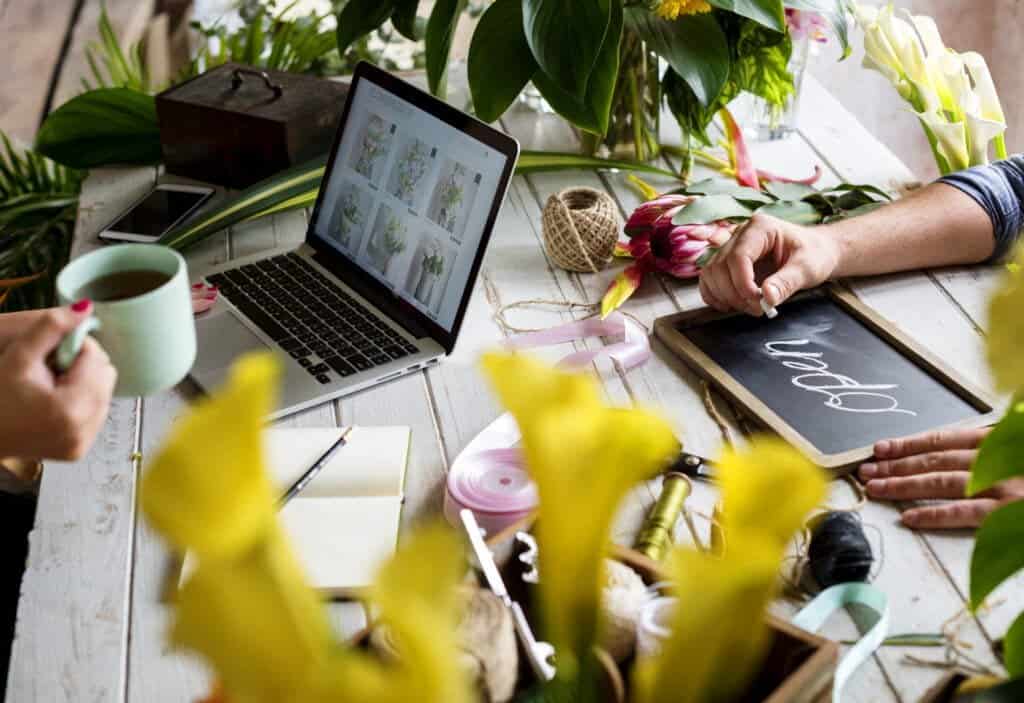 Creative flower arranging on a white wooden table with a laptop displaying floral images, a chalkboard sign with "Open," and surrounded by vibrant flowers and green plants.