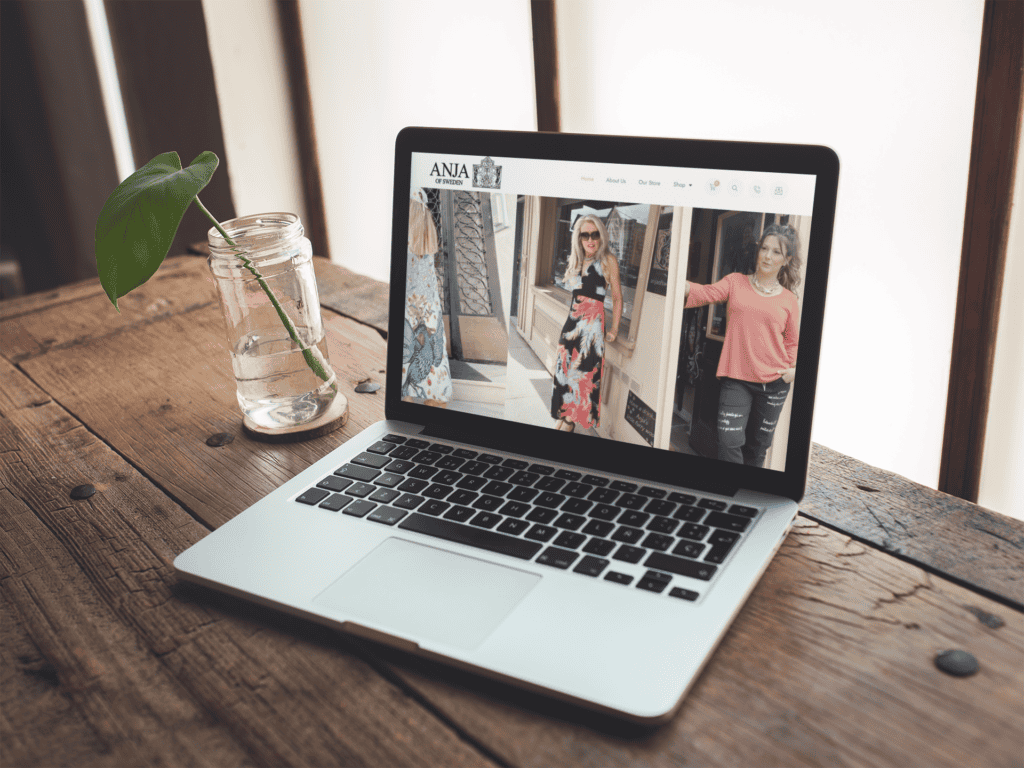 A laptop displaying an online fashion store, placed on a rustic wooden table with a glass jar containing a green leaf, in a cozy natural-lit setting.