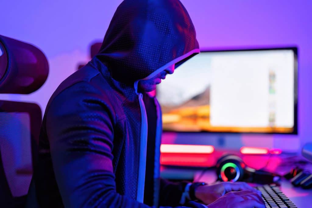 Hacker in a dark hoodie working on a computer with RGB lighting in a Tech-Savvy Home Office | Cybersecurity Threats and Digital Security Concept.
