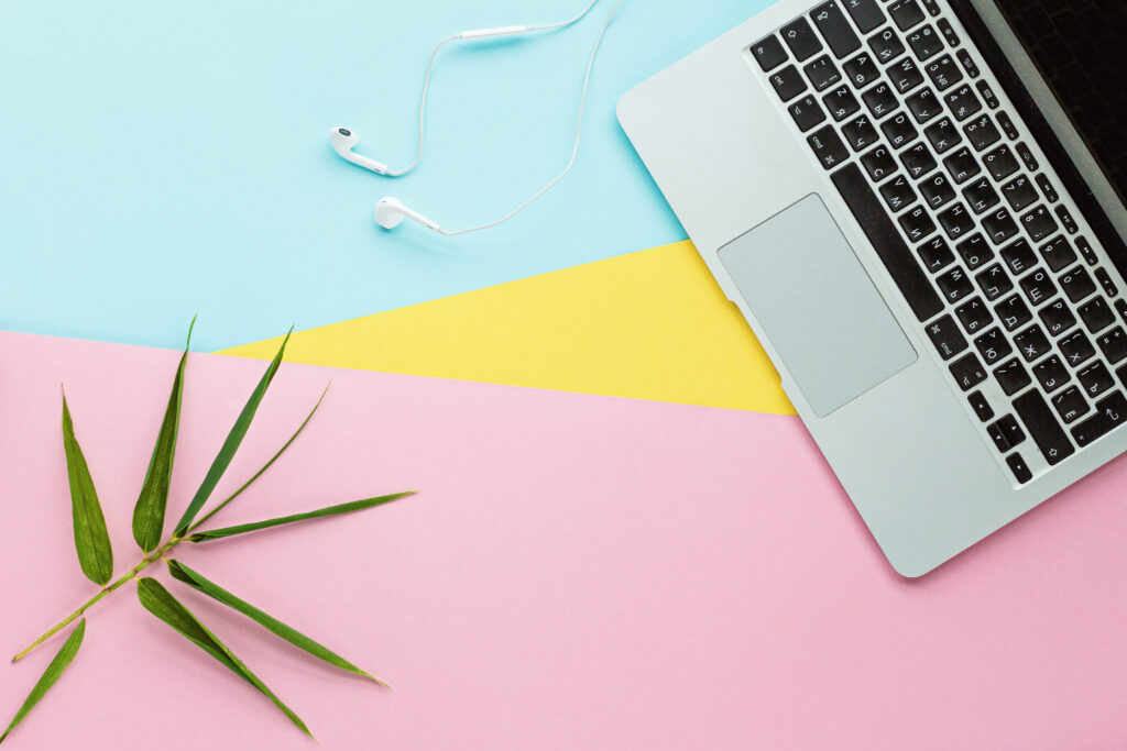 Laptop with keyboard and touchpad, white earphones, and a green plant on pastel pink, yellow, and blue background, suitable for web design and digital workspace themes.