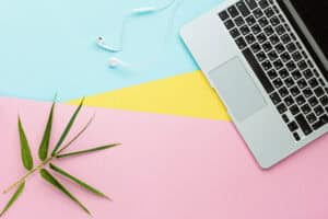 Laptop with keyboard and touchpad, white earphones, and a green plant on pastel pink, yellow, and blue background, suitable for web design and digital workspace themes.