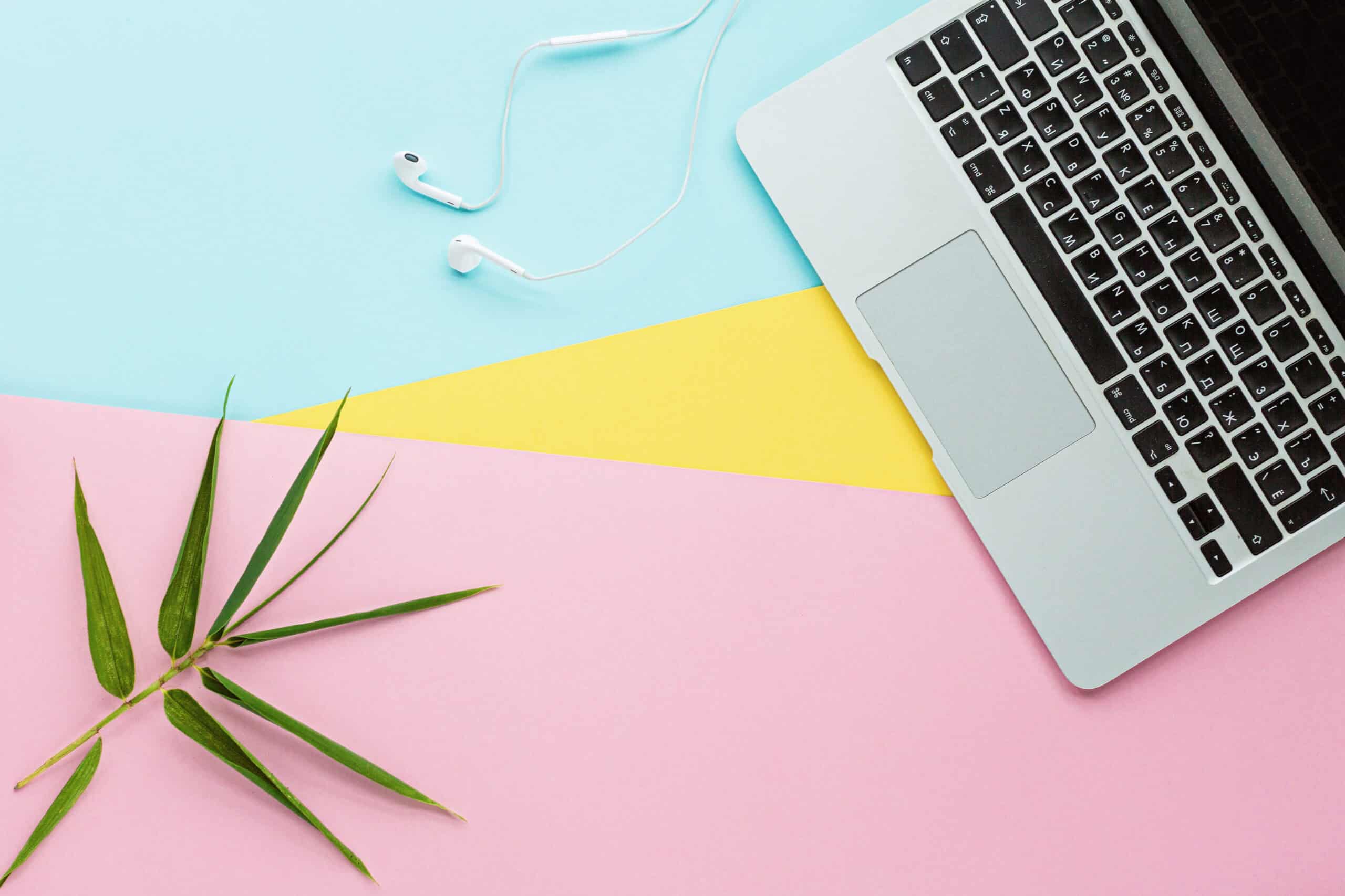 Laptop with keyboard and touchpad, white earphones, and a green plant on pastel pink, yellow, and blue background, suitable for web design and digital workspace themes.